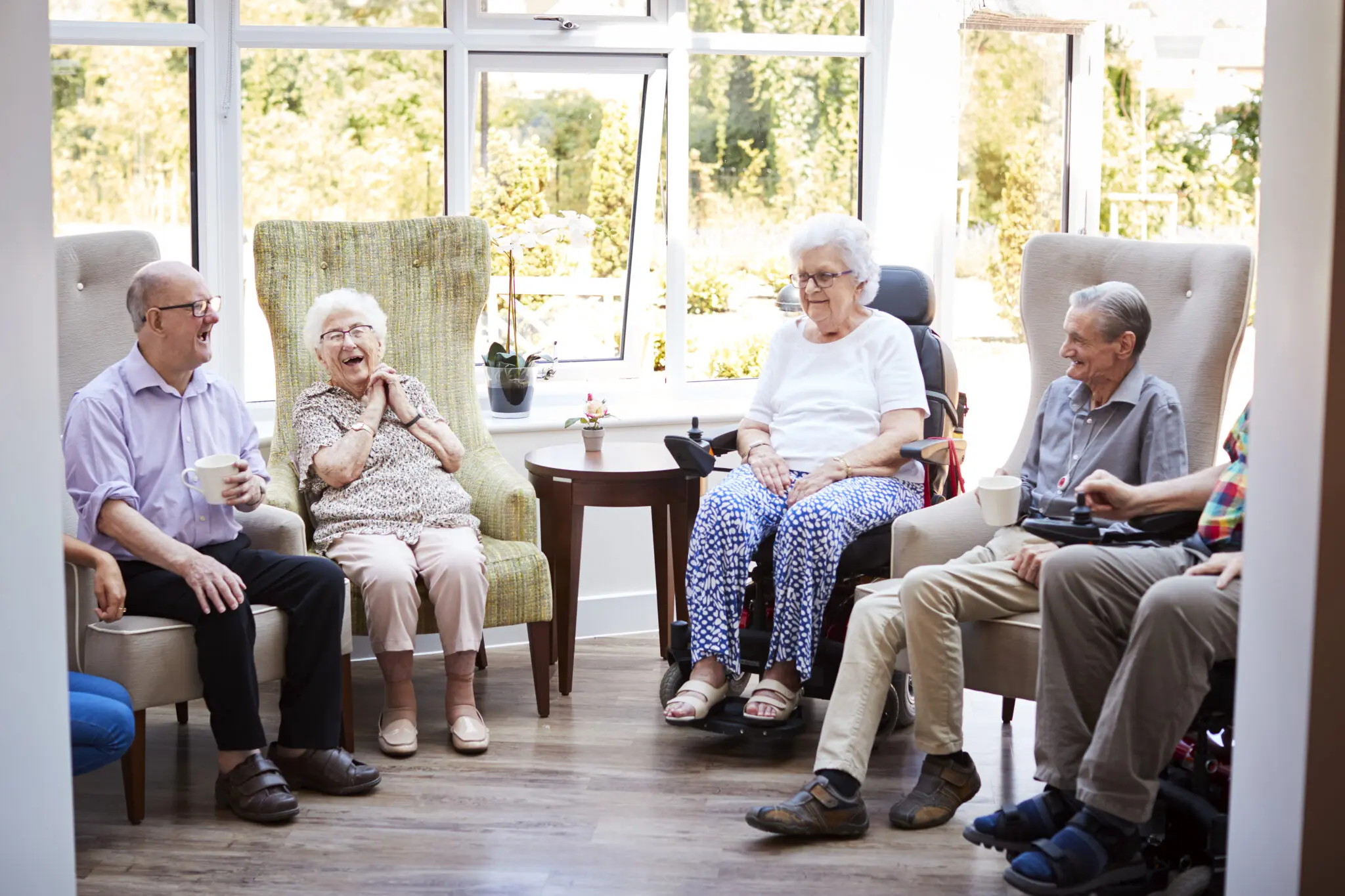 Male And Female Residents Sitting In Chairs And Talking In Lounge Of Retirement Home Male And Female Residents Sitting In Chairs And Talking In Lounge Of Retirement Home