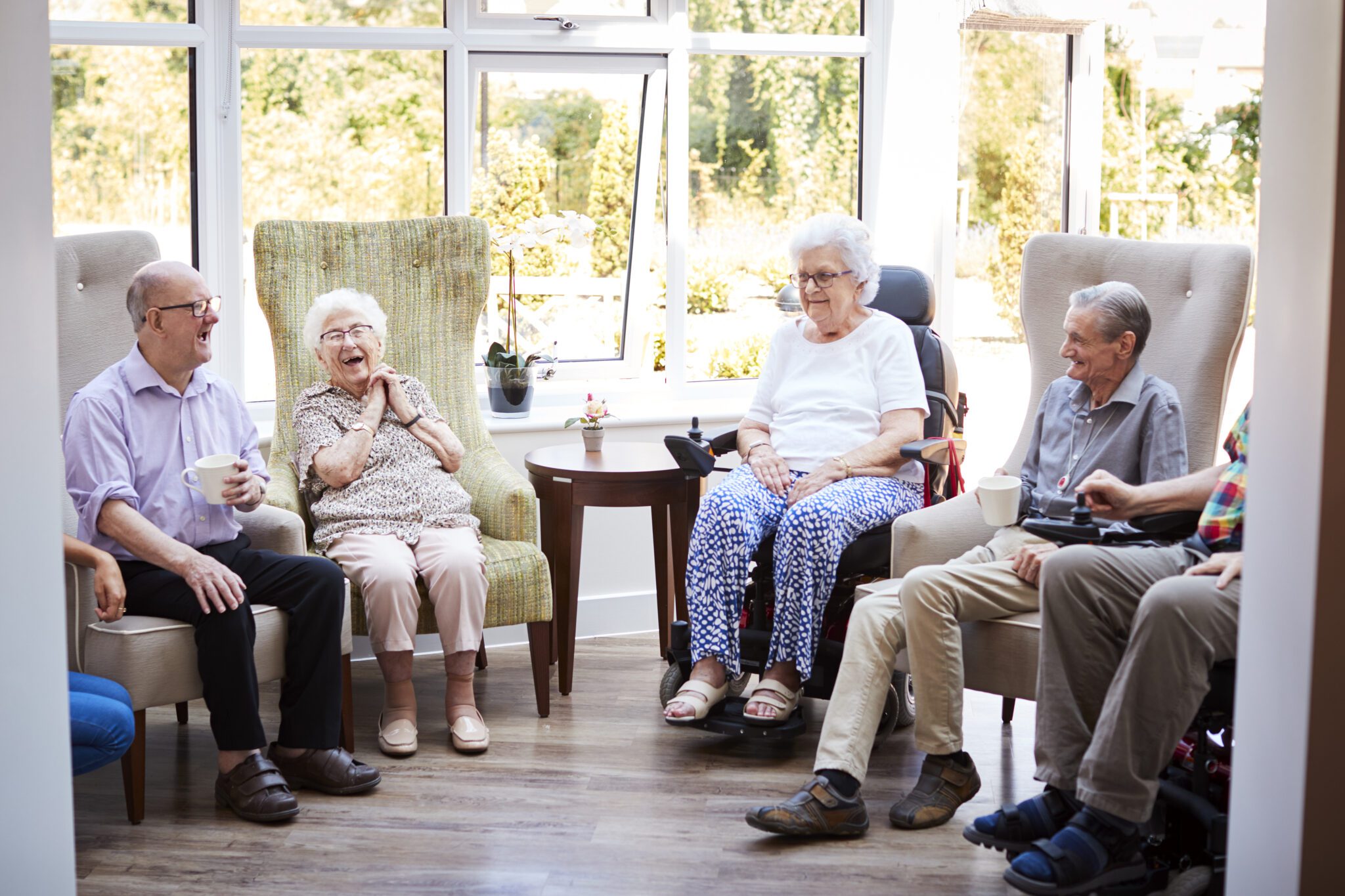 Male And Female Residents Sitting In Chairs And Talking In Lounge Of Retirement Home Male And Female Residents Sitting In Chairs And Talking In Lounge Of Retirement Home
