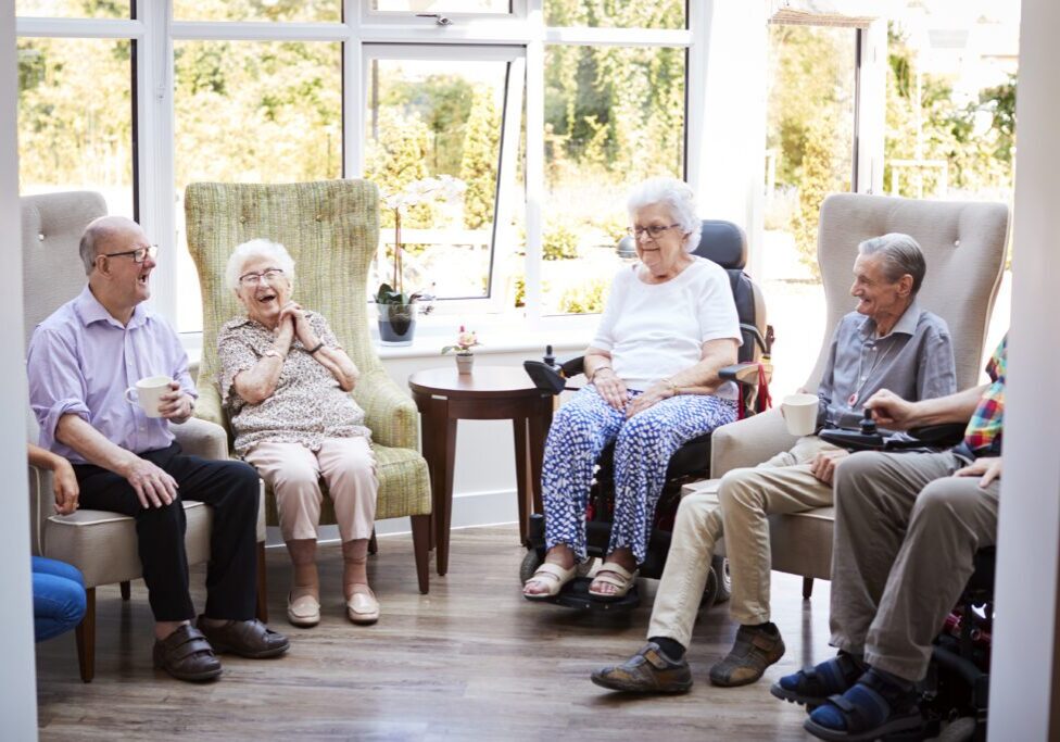 Male And Female Residents Sitting In Chairs And Talking In Lounge Of Retirement Home