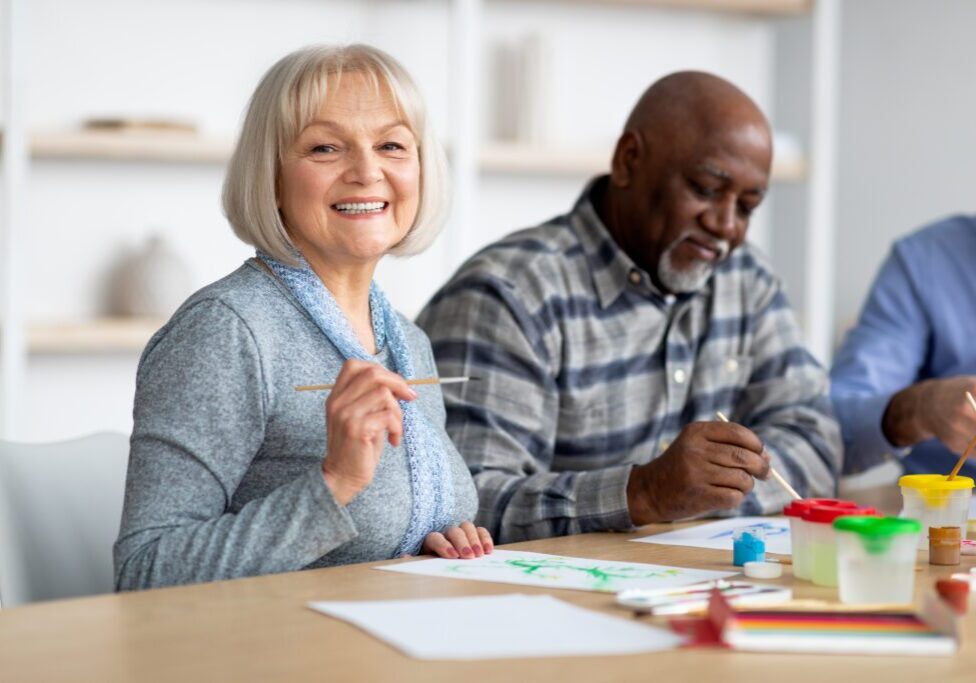 Art-therapy for seniors concept. Positive multiracial group of elderly people enjoying painting with brush, sitting around table, having good time together at assisted lliving, closeup photo